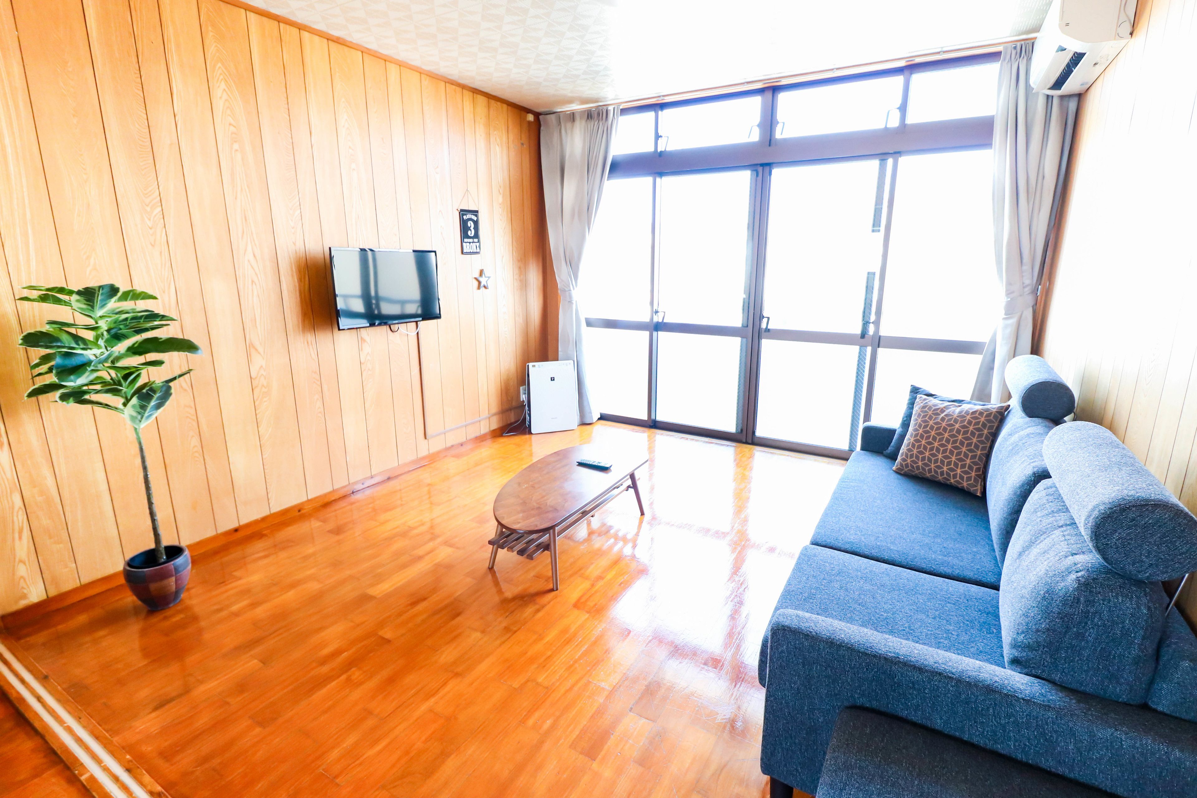 Ein Zimmer mit Holzboden, einem blauen Sofa, einem kleinen Tisch, einem Fernseher und einem großen Fenster mit Vorhängen.