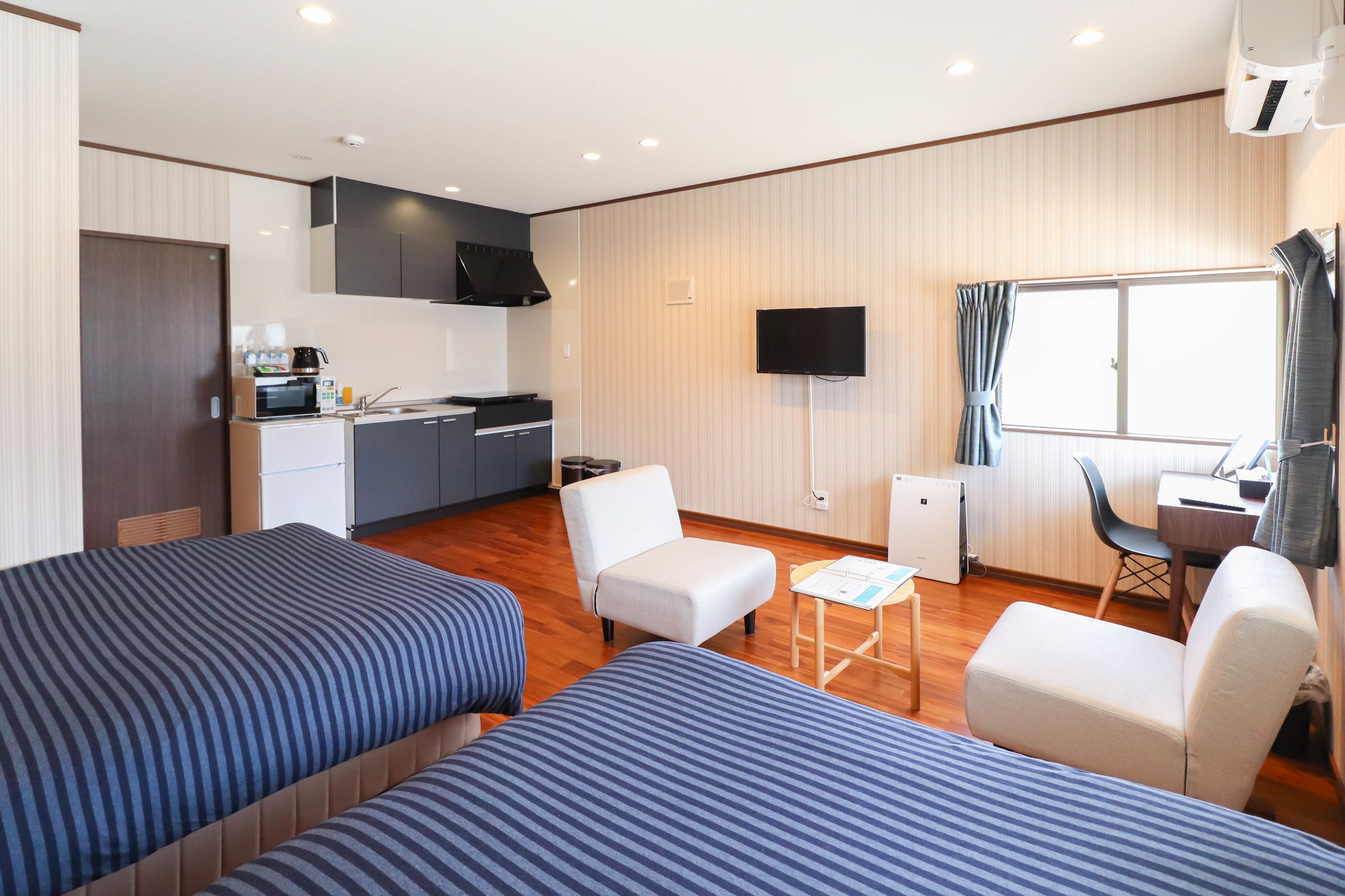 A hotel room with two beds, a kitchenette, a TV, and a window with curtains.