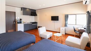 A hotel room with two beds, a kitchenette, a TV, and a window with curtains.
