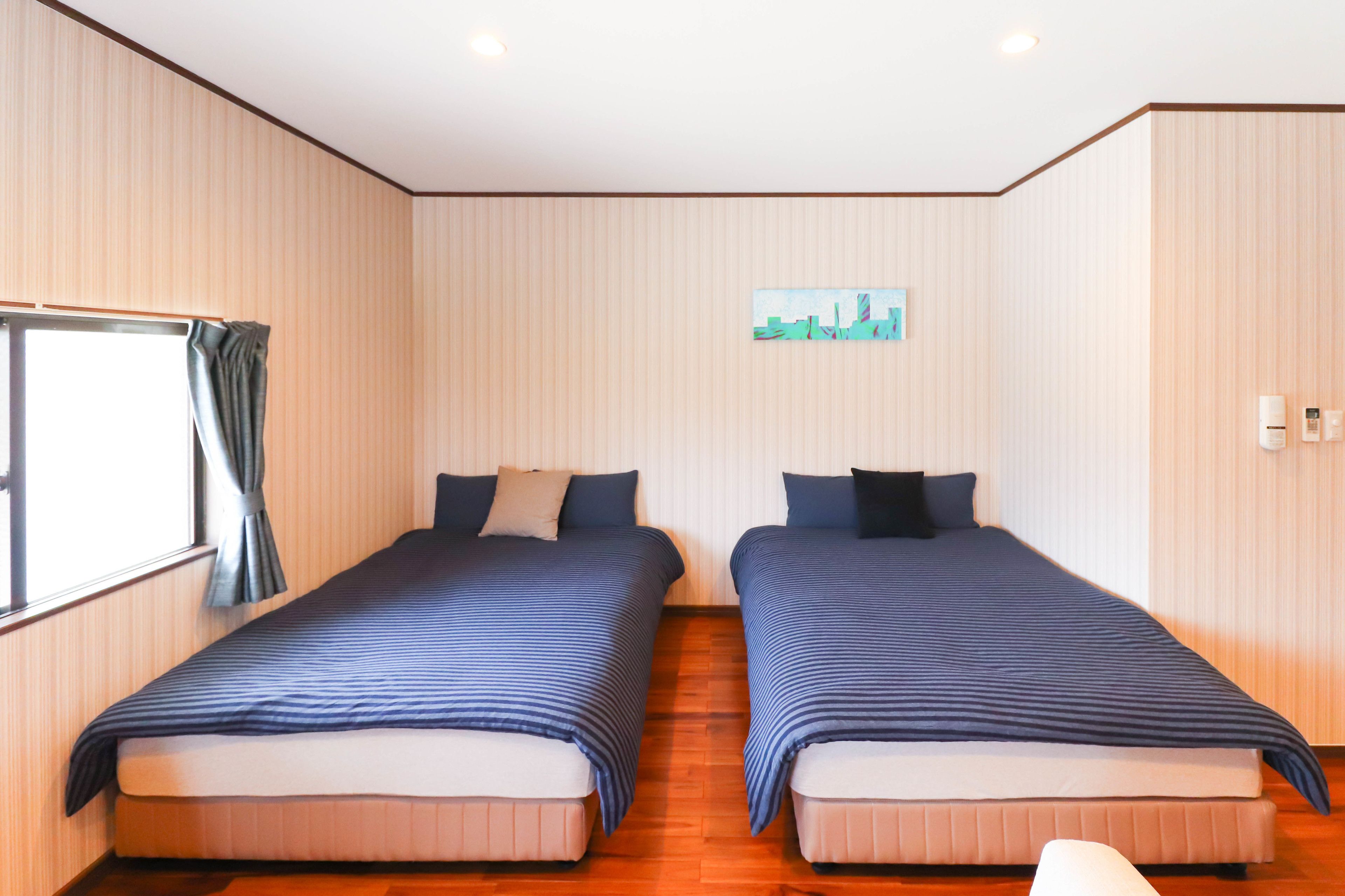 A room with two beds, wooden flooring, a window with curtains, and a small painting on the wall.