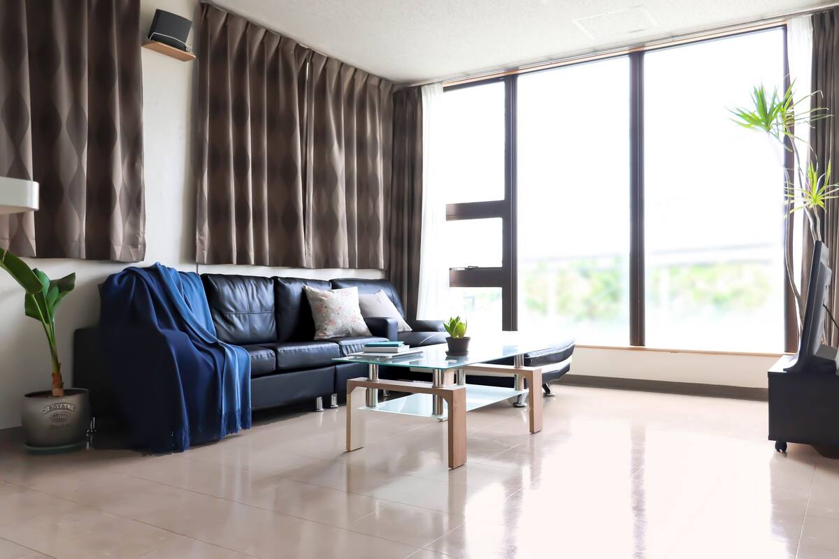 A modern living room with a leather sofa, a glass coffee table, and a large window with curtains.