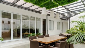 An outdoor dining area with a wooden table and wicker chairs, surrounded by potted plants and a glass door leading to a kitchen area. - RedDoorz Plus near Exit Toll Puncak 2 (Ciawi)