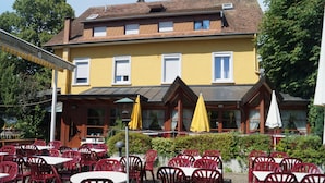 Un edificio giallo con tetto in tegole, sedute all'aperto con sedie rosse e tavoli bianchi, e ombrelloni gialli.