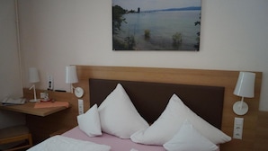 Camera d'albergo con un letto, cuscini, un comodino con una lampada, una scrivania con una lampada e un quadro raffigurante un lago appeso al muro.