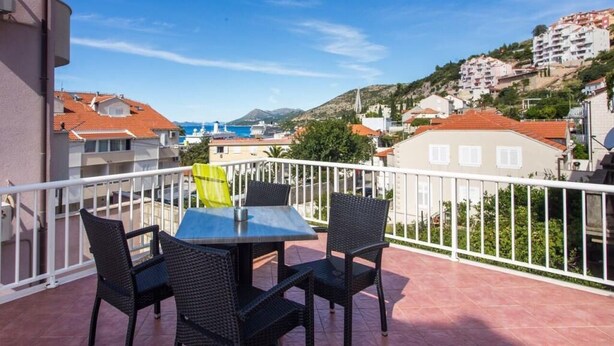 En balkon med bord og stole, udsigt over en kystby med bygninger og hav.