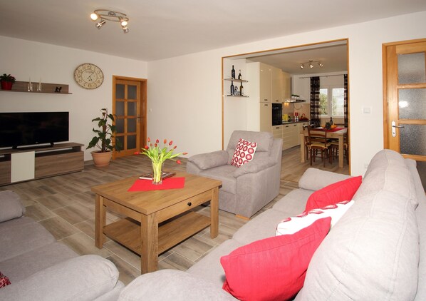 A living room with a TV, sofa, coffee table, and a dining area in the background.