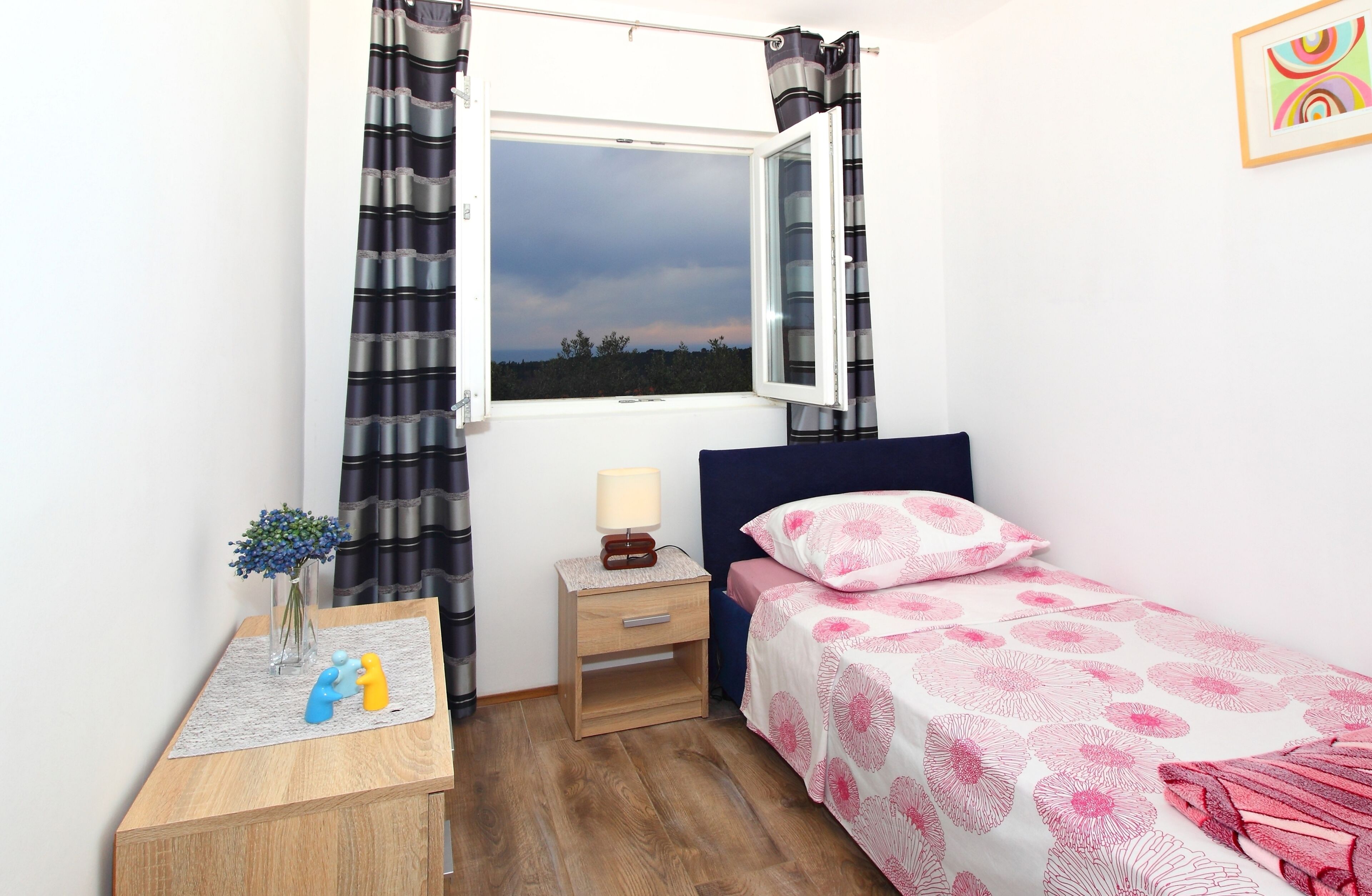 A bedroom with a single bed, a nightstand with a lamp, a small table with a vase and figurines, and a window with curtains.