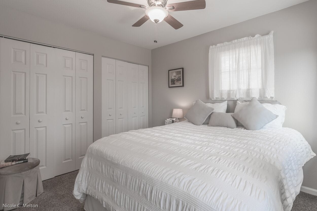 A bedroom with a large bed, a ceiling fan, white walls, and a window with sheer curtains.
