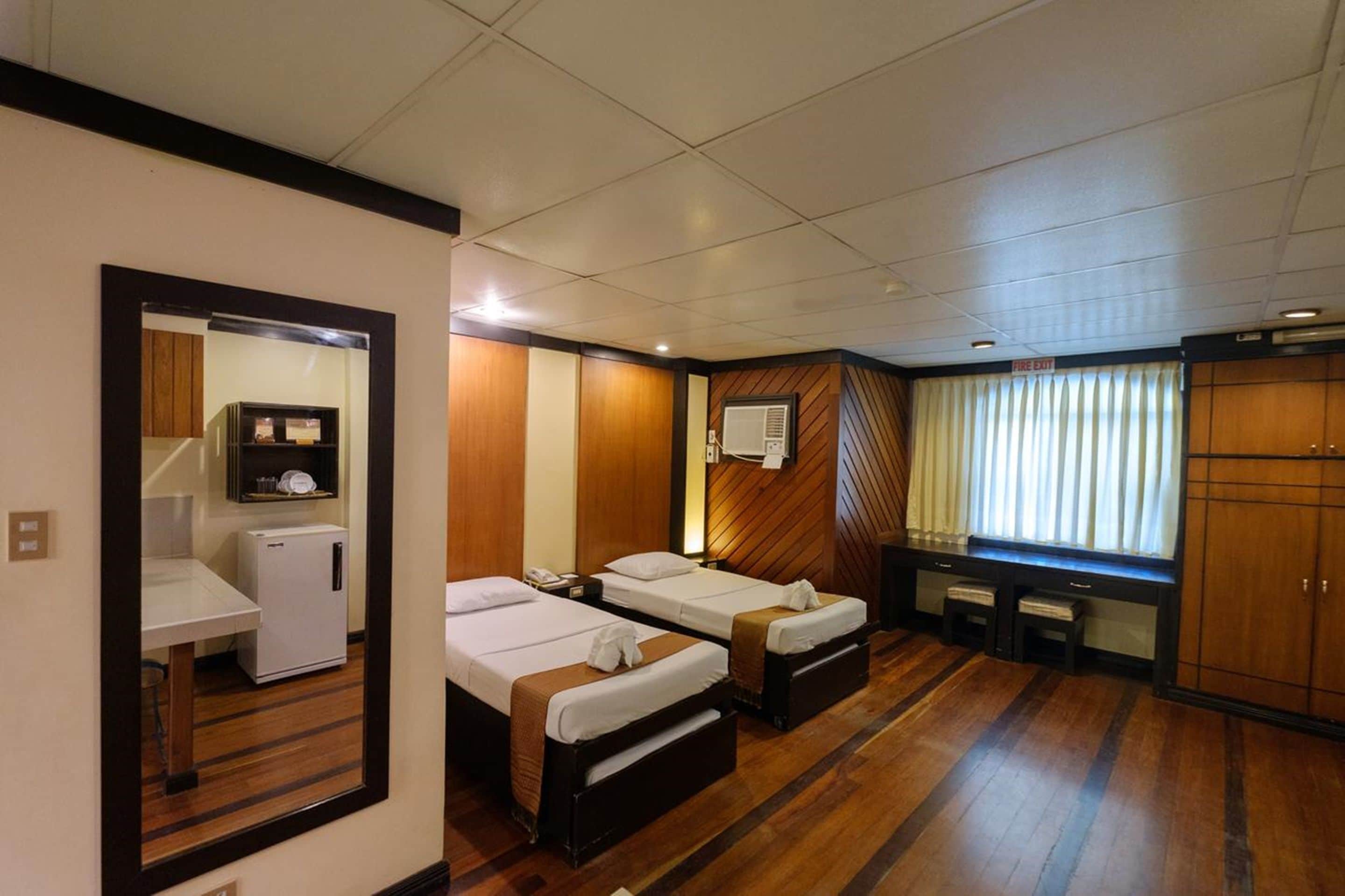 A hotel room with two single beds, a small desk with a refrigerator, a mirror, and a wardrobe.