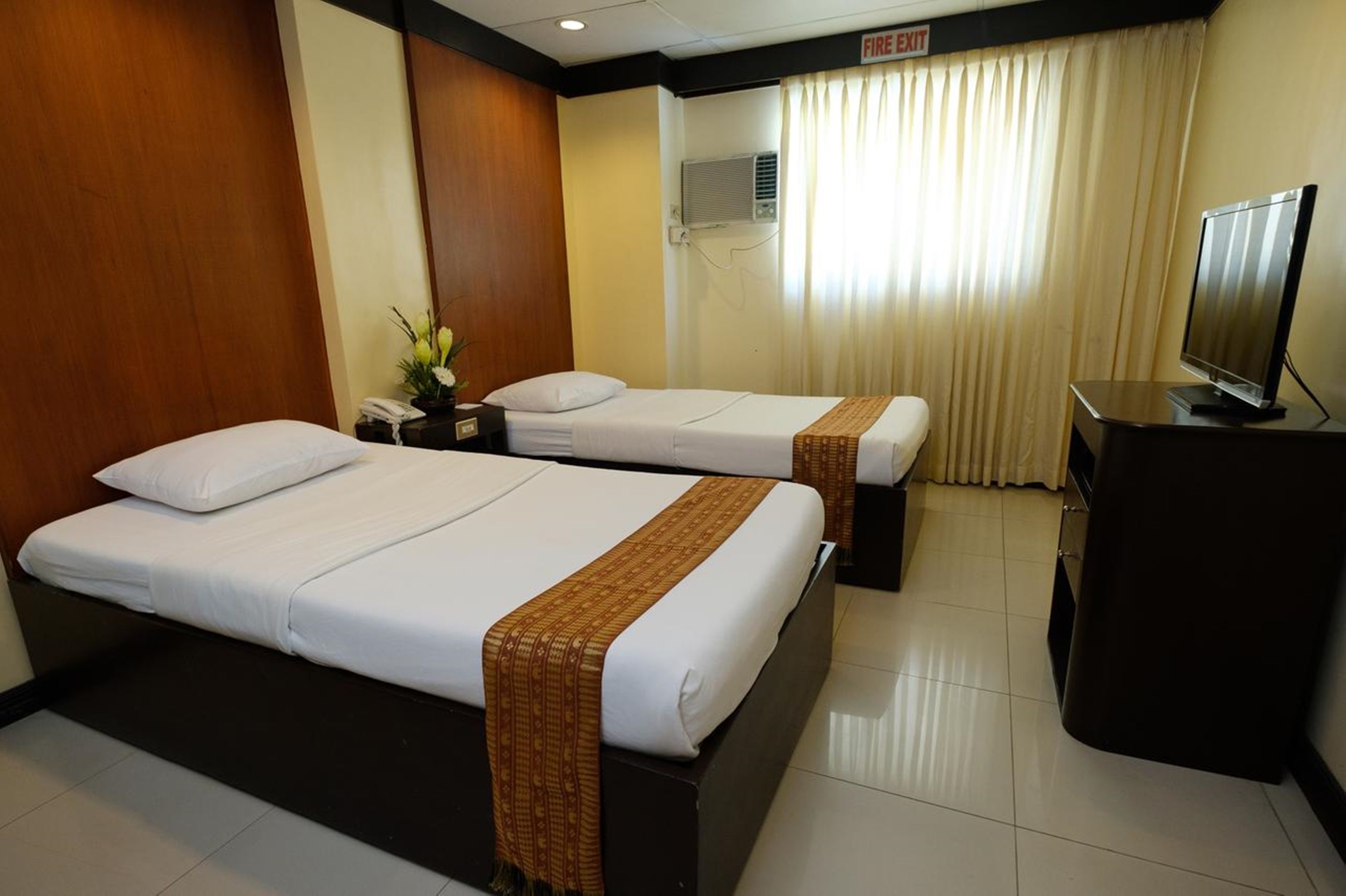 A hotel room with two beds, a television, a desk, and a fire exit sign.