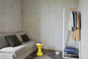 A room with a grey sofa, a yellow side table, a white shoe rack, and a grey floor. - Combo Milano - Hostel (Milan)