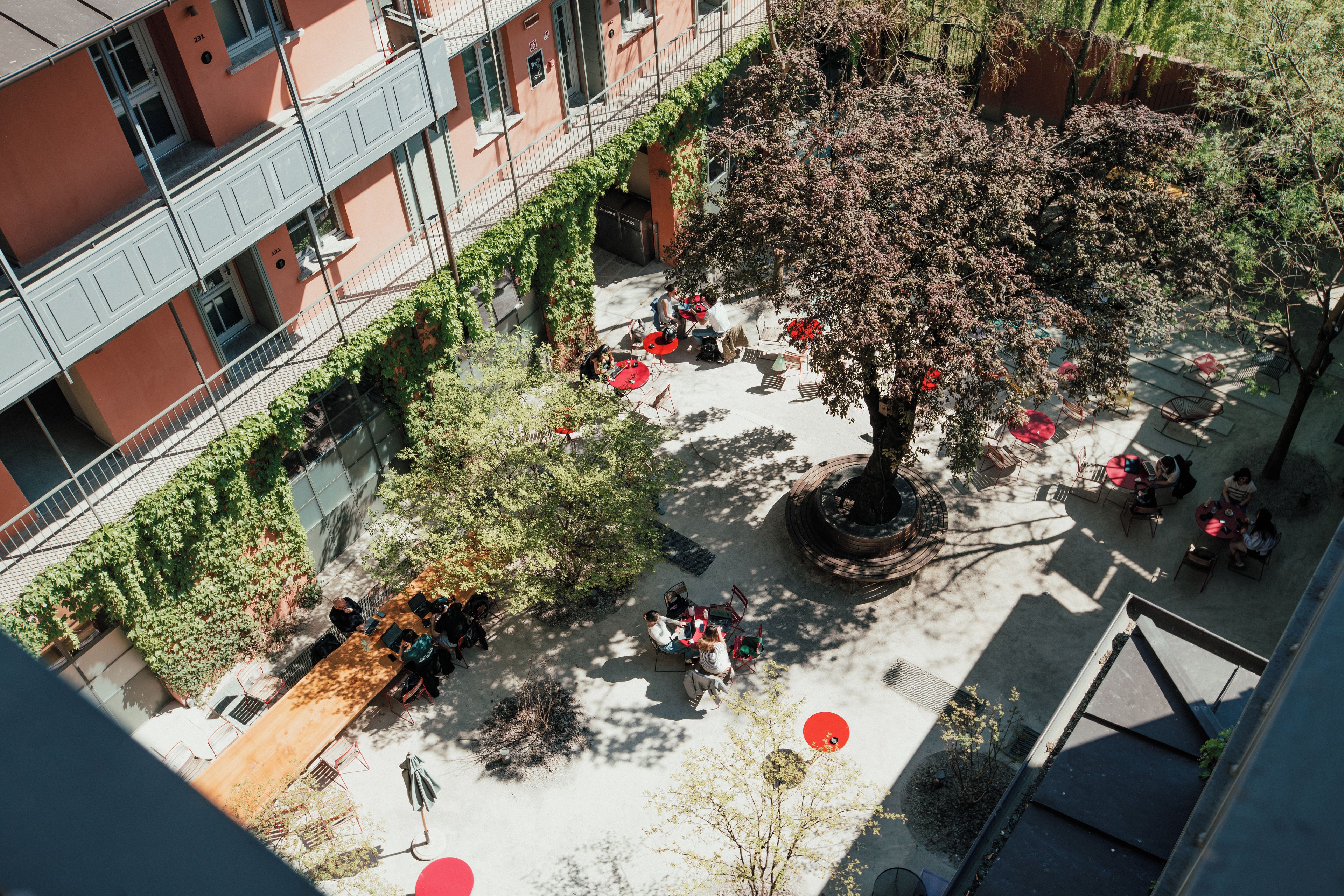 Ein Innenhof mit Sitzbereichen, einem zentralen Baum und einem Gebäude mit Balkonen.