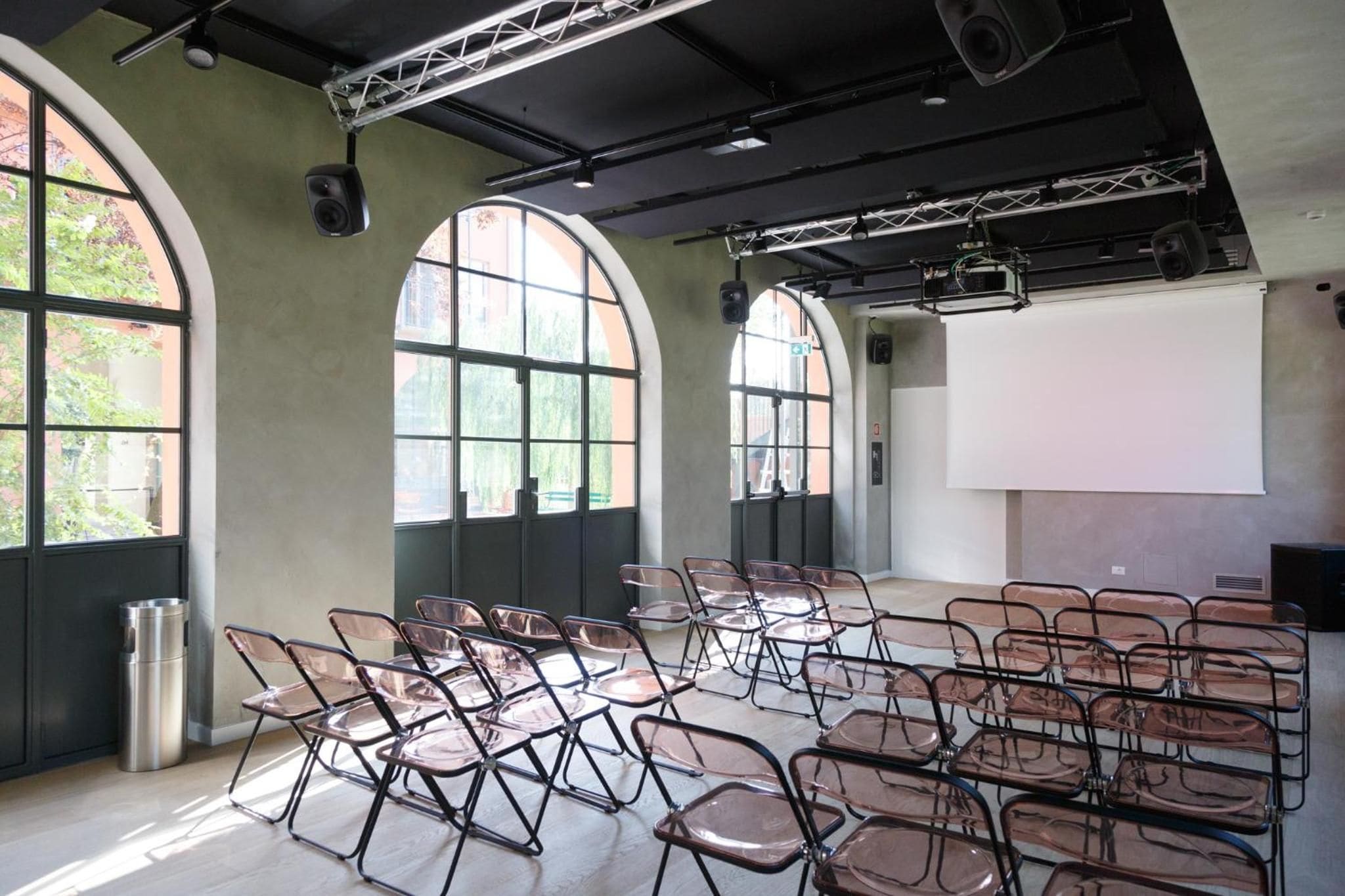 A conference room with arched windows, a white projection screen, and rows of foldable chairs.