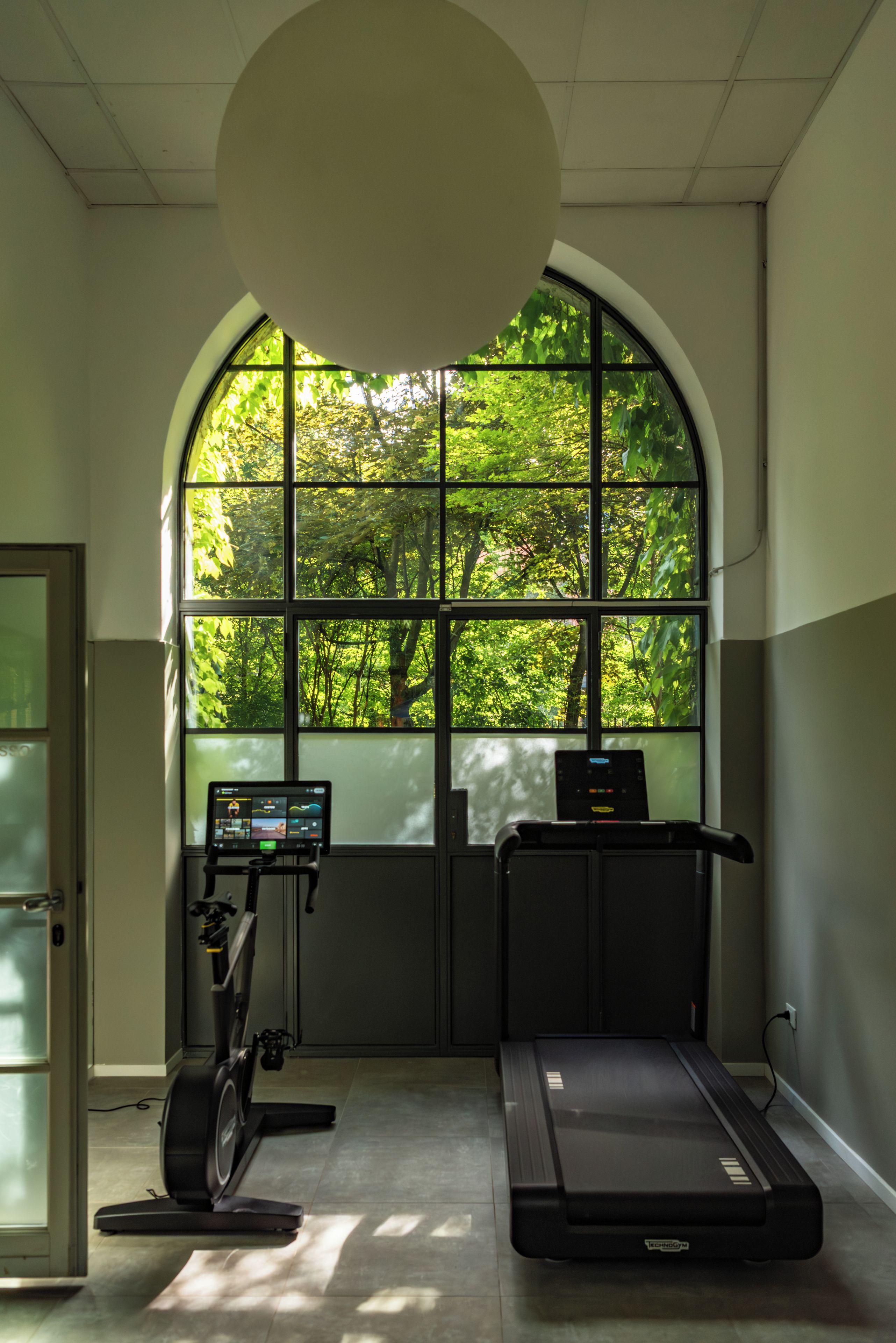 Ein Fitnessstudio mit Laufband, Ergometer und einem großen Bogenfenster mit Blick auf eine grüne Landschaft.