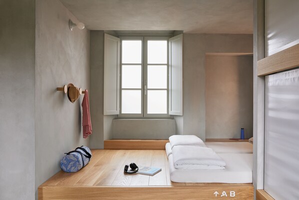 A minimalist room with a wooden floor, a window, a bed, a bench, and a yoga mat. - Combo Milano - Hostel (Milan)