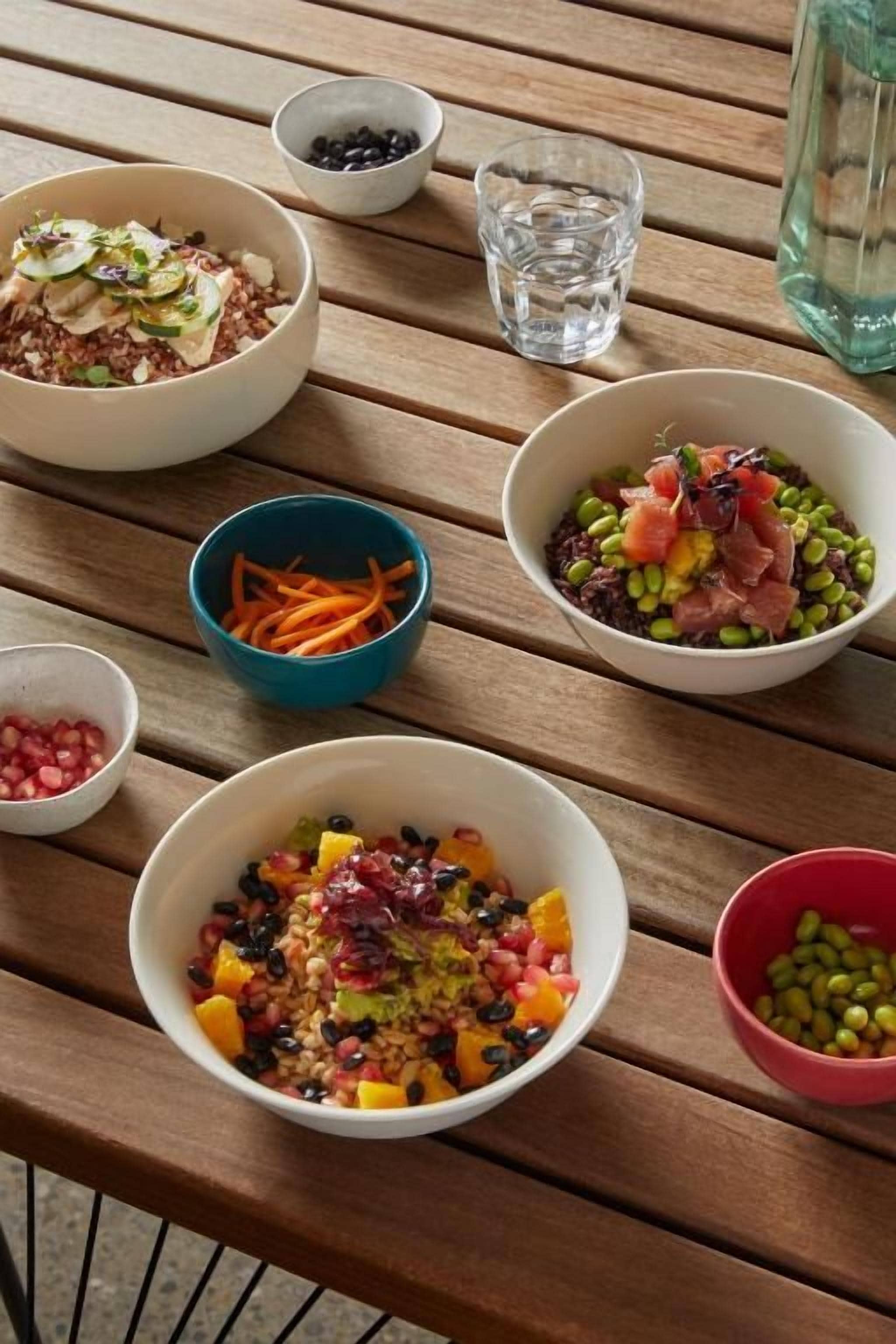 Una mesa de madera con varios recipientes que contienen diferentes alimentos, un vaso de agua y una botella.