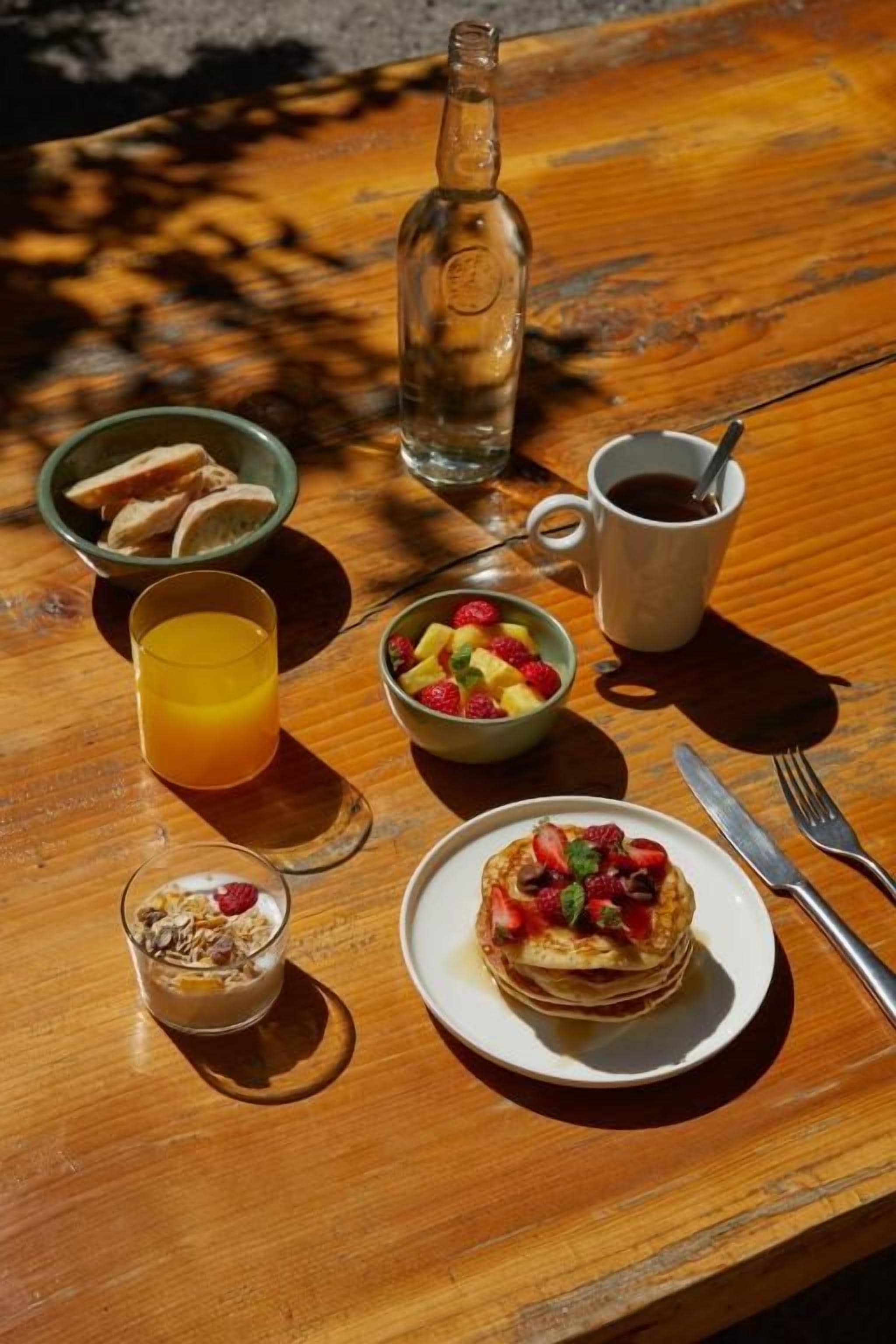 Una mesa de madera con productos para el desayuno, incluyendo tortitas, un bol de fruta, un vaso de zumo de naranja, una botella de agua y una taza de café.