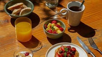 Una mesa de madera con productos para el desayuno, incluyendo tortitas, un bol de fruta, un vaso de zumo de naranja, una botella de agua y una taza de café.