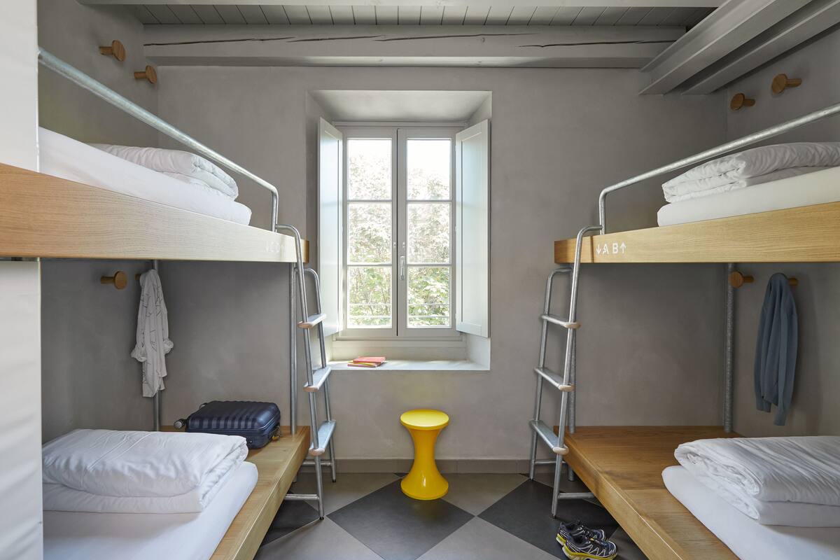 Un dormitorio con literas, una ventana pequeña, un taburete amarillo y una maleta.