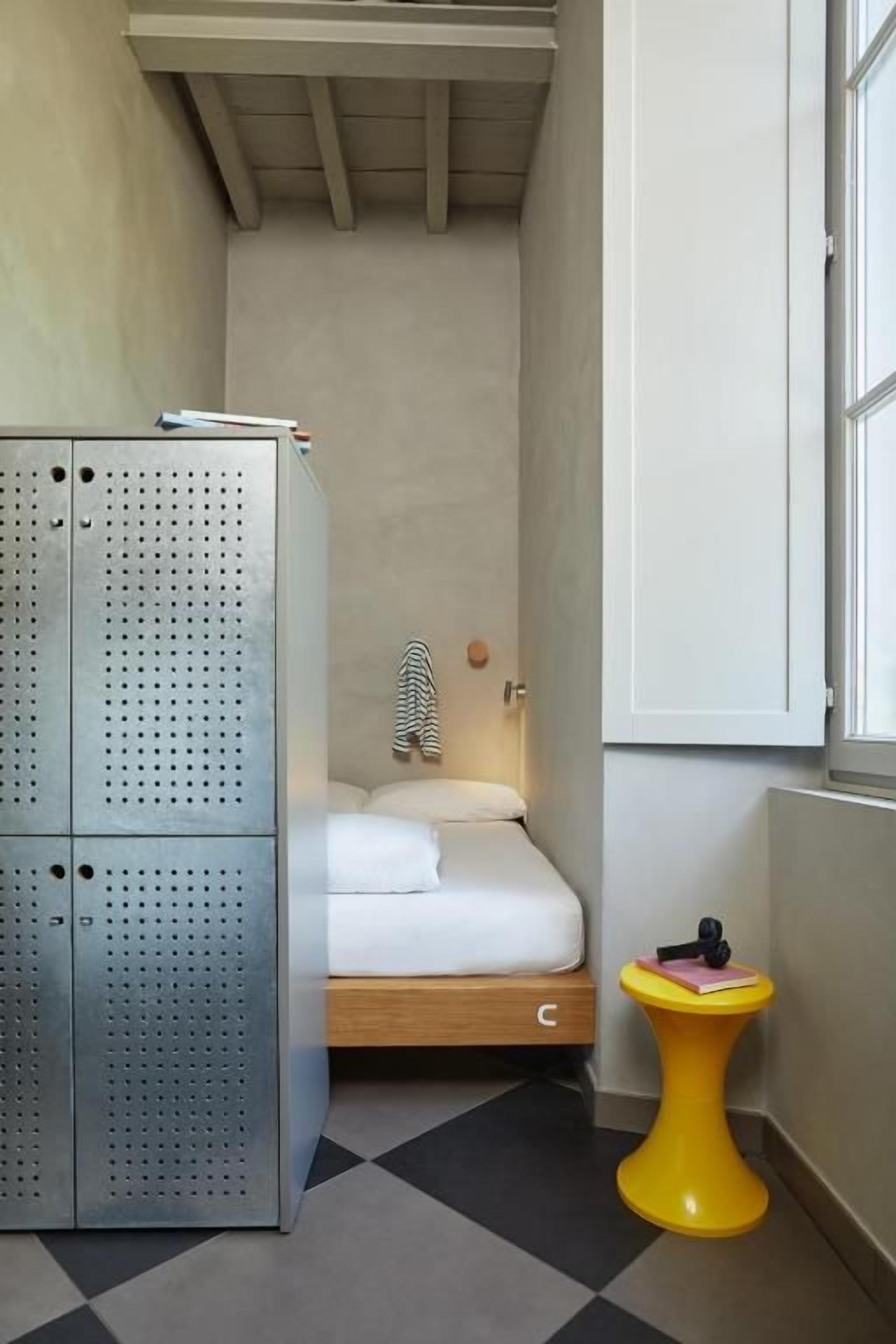 A compact room with a bed, a small yellow stool, and a metal wardrobe.