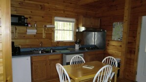 A compact kitchen with wooden cabinets, a stainless steel stove, a microwave, and a sink. There is a dining table with four chairs. - Acadia Explorer Cabin Rentals Cabin 1  (Southwest Harbor)