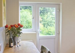 A table with a bottle of wine, a wine glass, and a vase with roses by a window with a view of greenery.