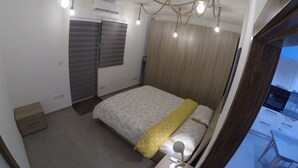 Een slaapkamer met een bed, nachtkastje, lamp en uitzicht op de keuken.