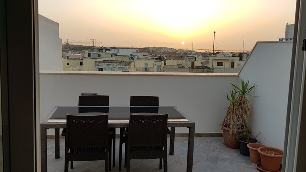 Een balkon met een tafel en stoelen, met uitzicht op een stadsgezicht bij zonsondergang.