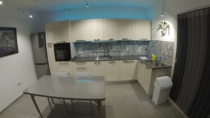 Een moderne keuken met lichte kastjes, een roestvrijstalen aanrechtblad en een ingebouwde oven. In het midden staat een tafel met stoelen, en rechts een prullenbak.