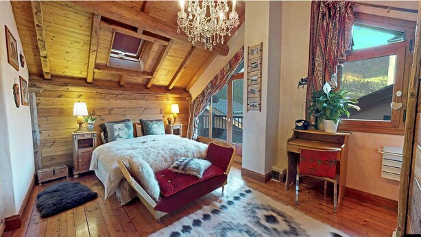 A cozy bedroom with a sloped ceiling, a chandelier, a bed with a red headboard, a nightstand with a lamp, a window with curtains, and a plant on a side table.
