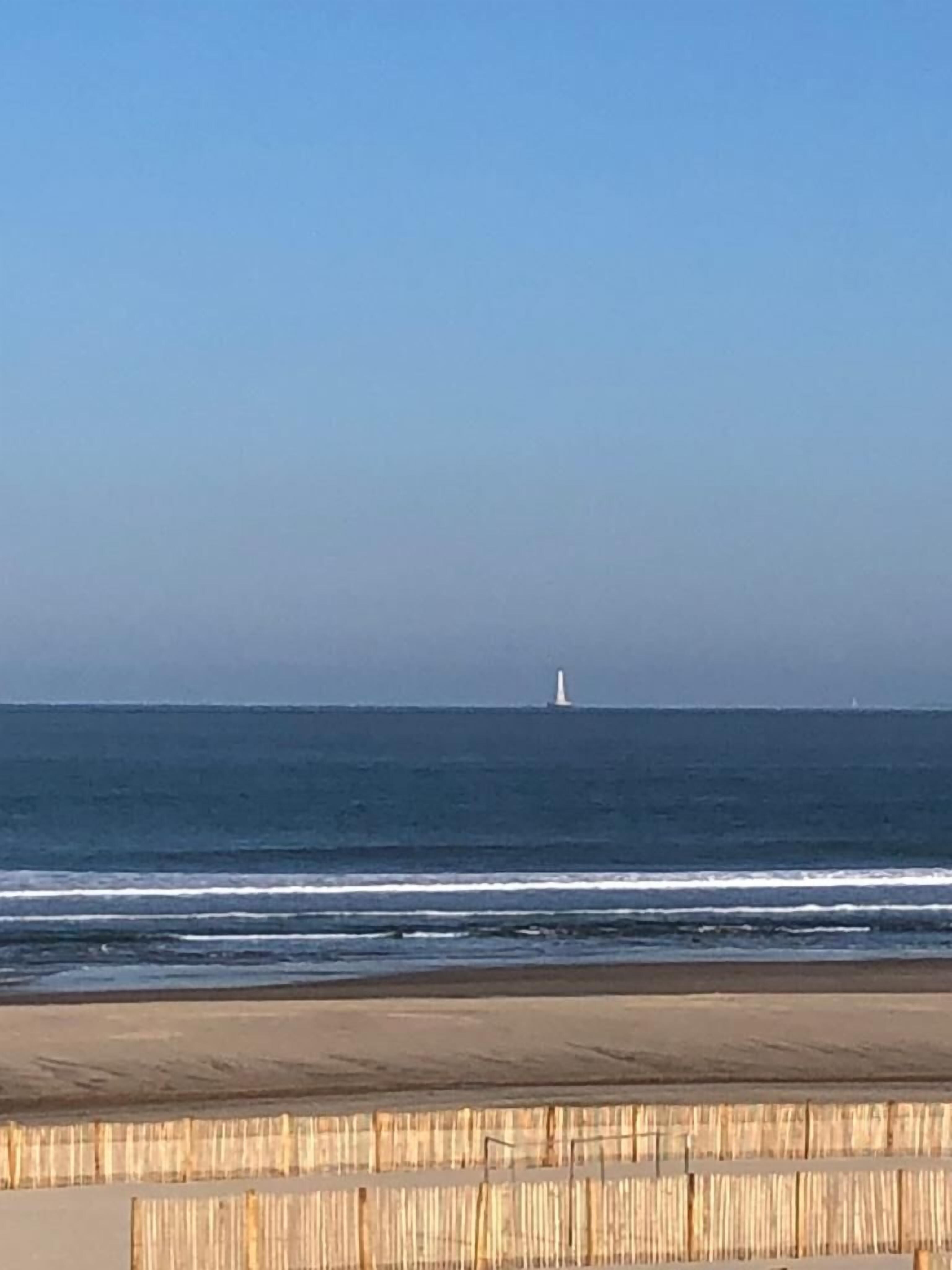 A beach with a lighthouse in the distance, calm sea, and a wooden fence in the foreground.