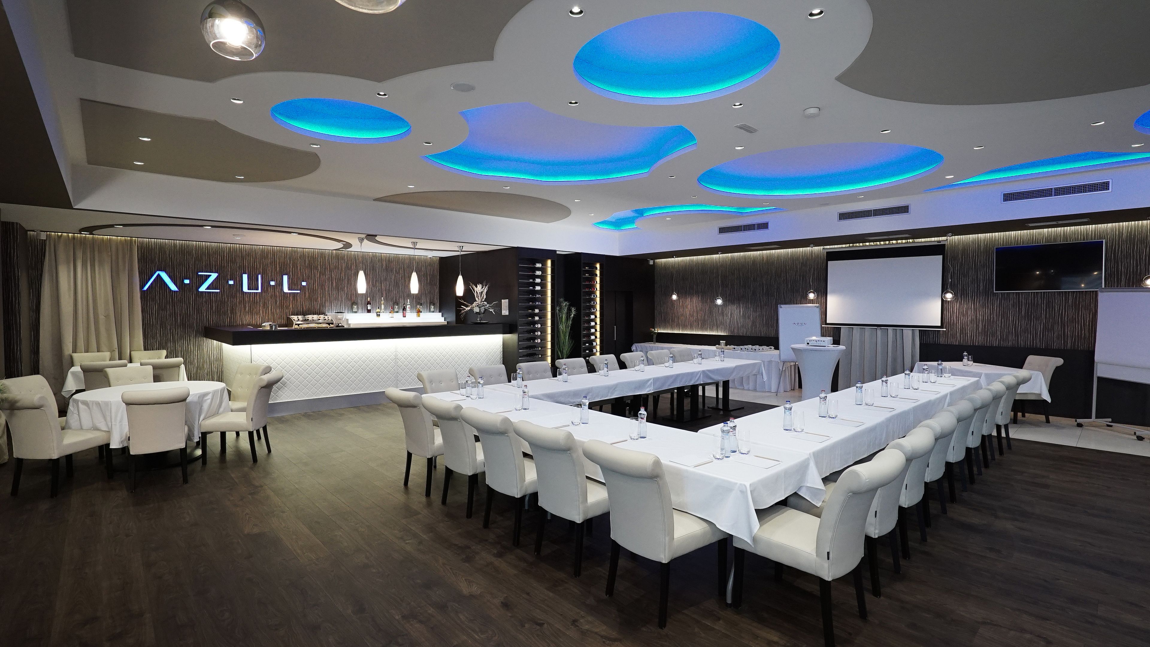 Une salle de conférence moderne, dotée d’une longue table, de chaises blanches, d’un coin bar et d’un grand écran.