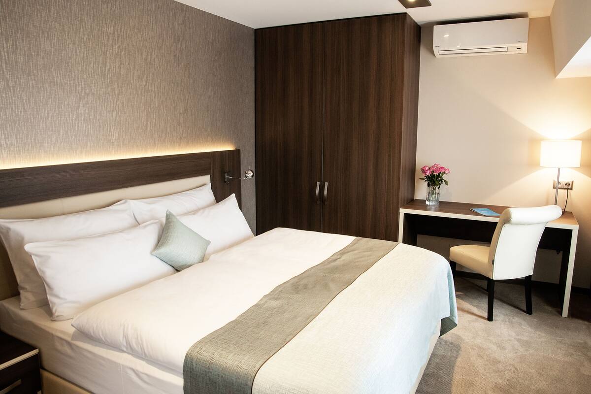 Hotellihuoneessa on suuri sänky, työpöytä ja tuoli. Seinällä on ilmastointilaite ja työpöydällä lamppu.