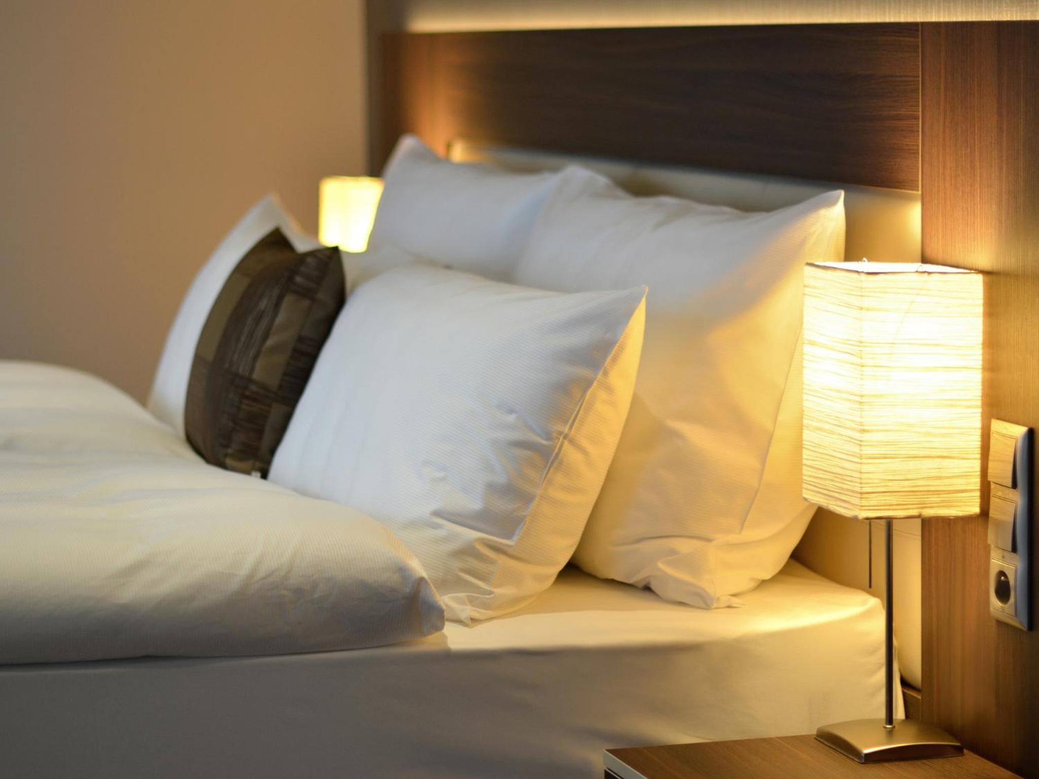A neatly made bed with white linens and pillows, a bedside lamp, and a wooden headboard.