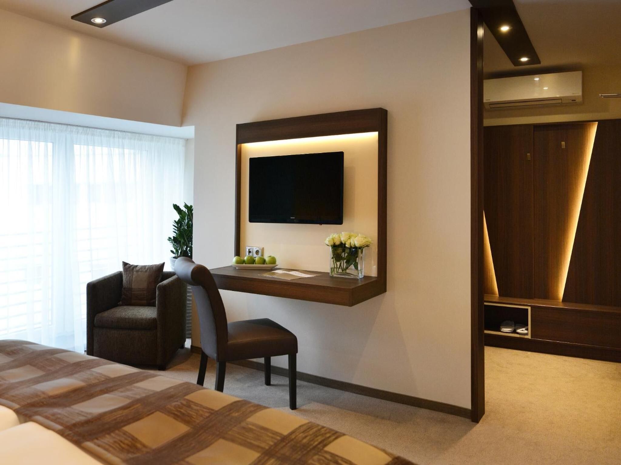 A hotel room with a bed, a desk with a chair, a TV mounted on the wall, and a wardrobe.