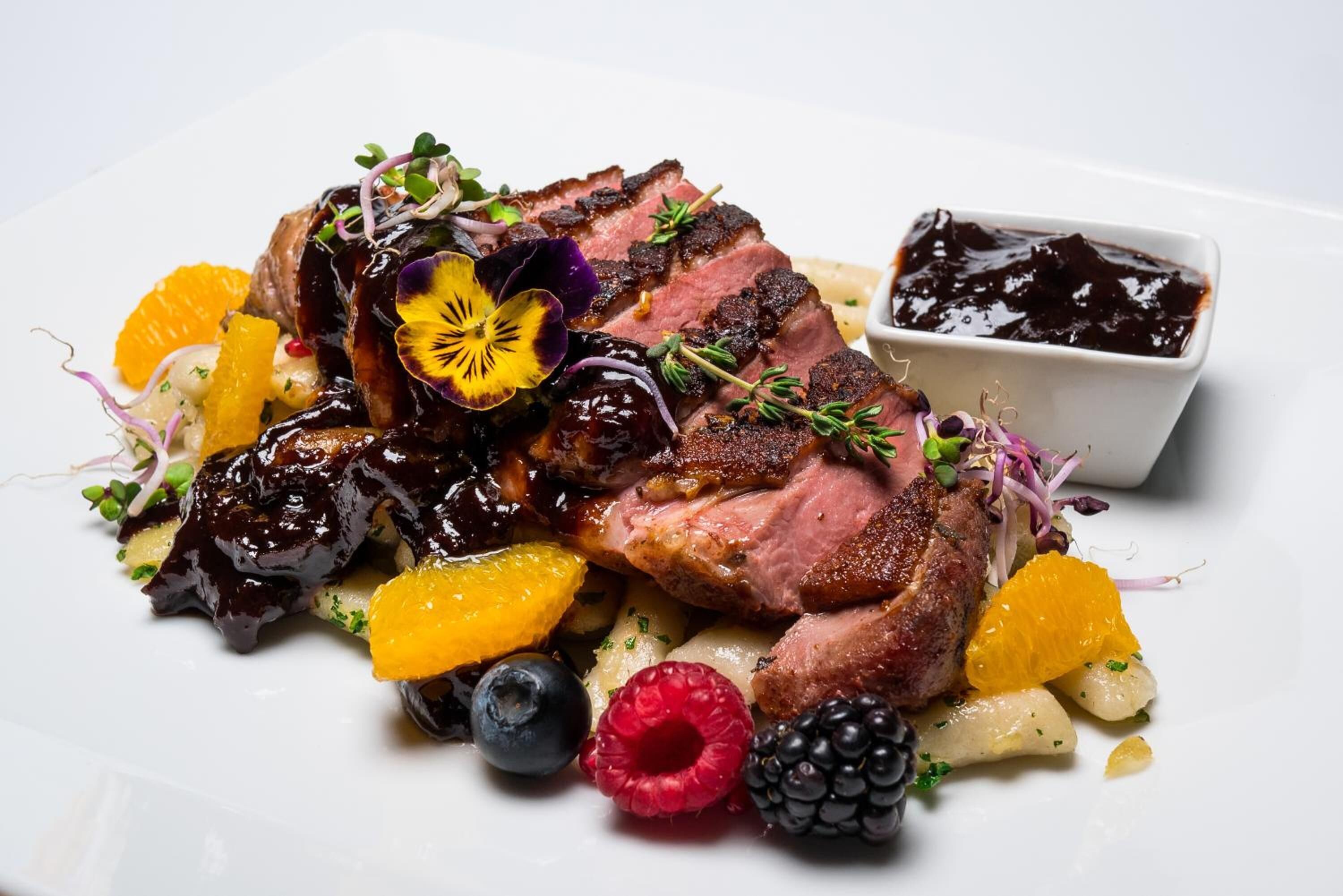 A plate of food with sliced meat, fruits, and a small bowl of sauce.