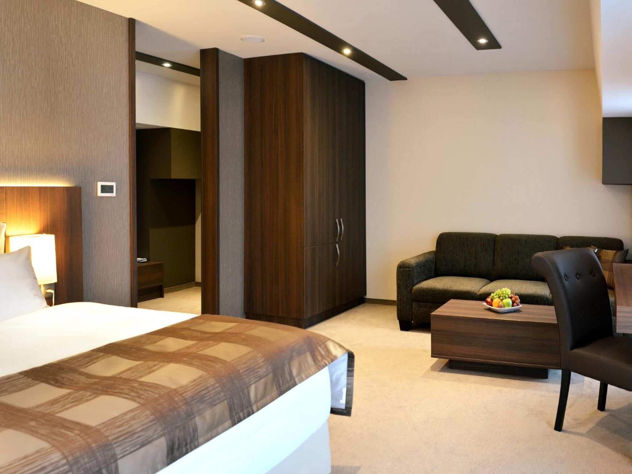 Een moderne hotelkamer met een groot bed, een donkere houten garderobe, een sofa en een klein tafeltje met fruit.