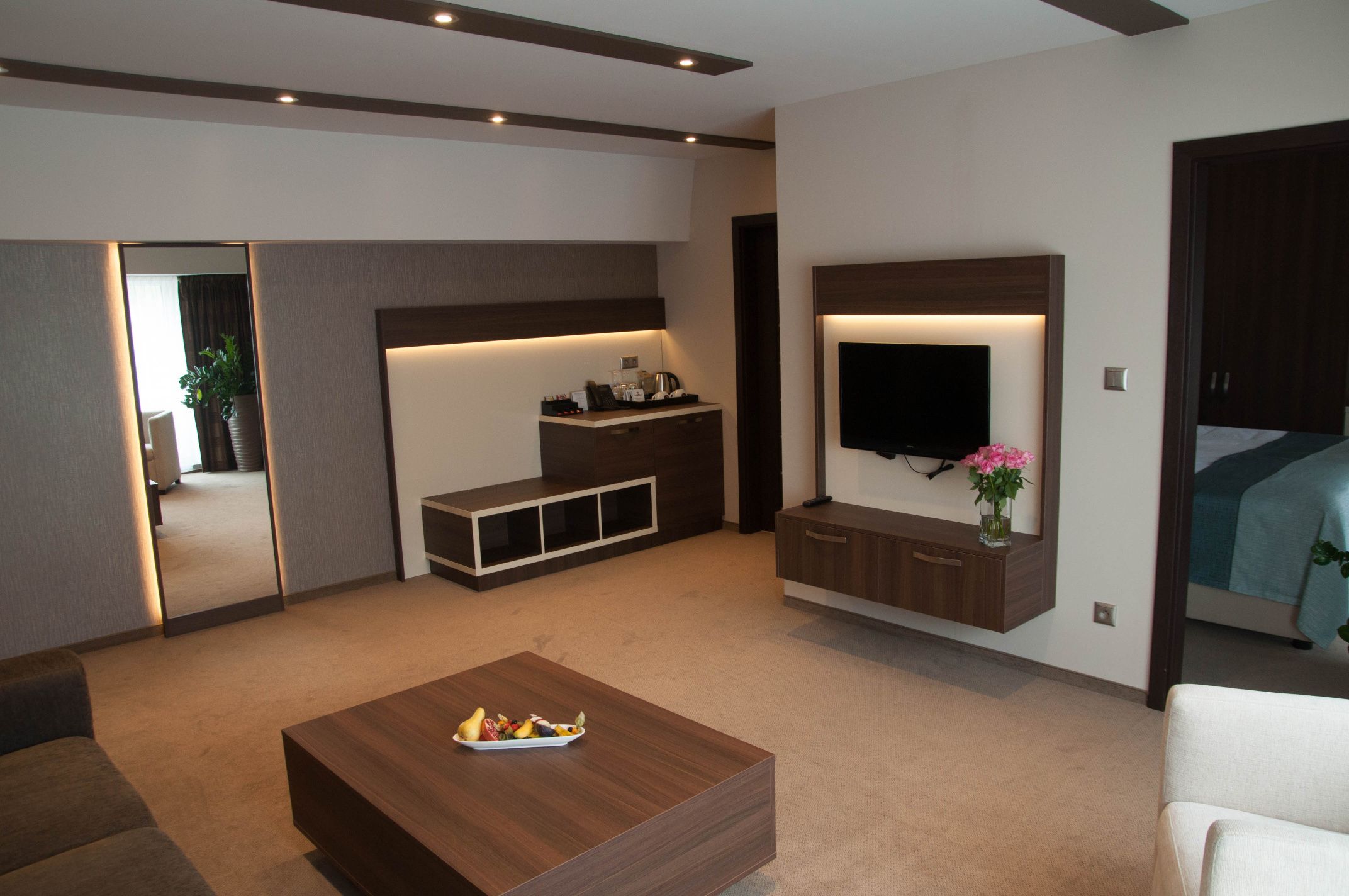 A modern living room with a flat-screen TV, a coffee table with fruit, a sofa, and a TV stand with a vase of flowers.
