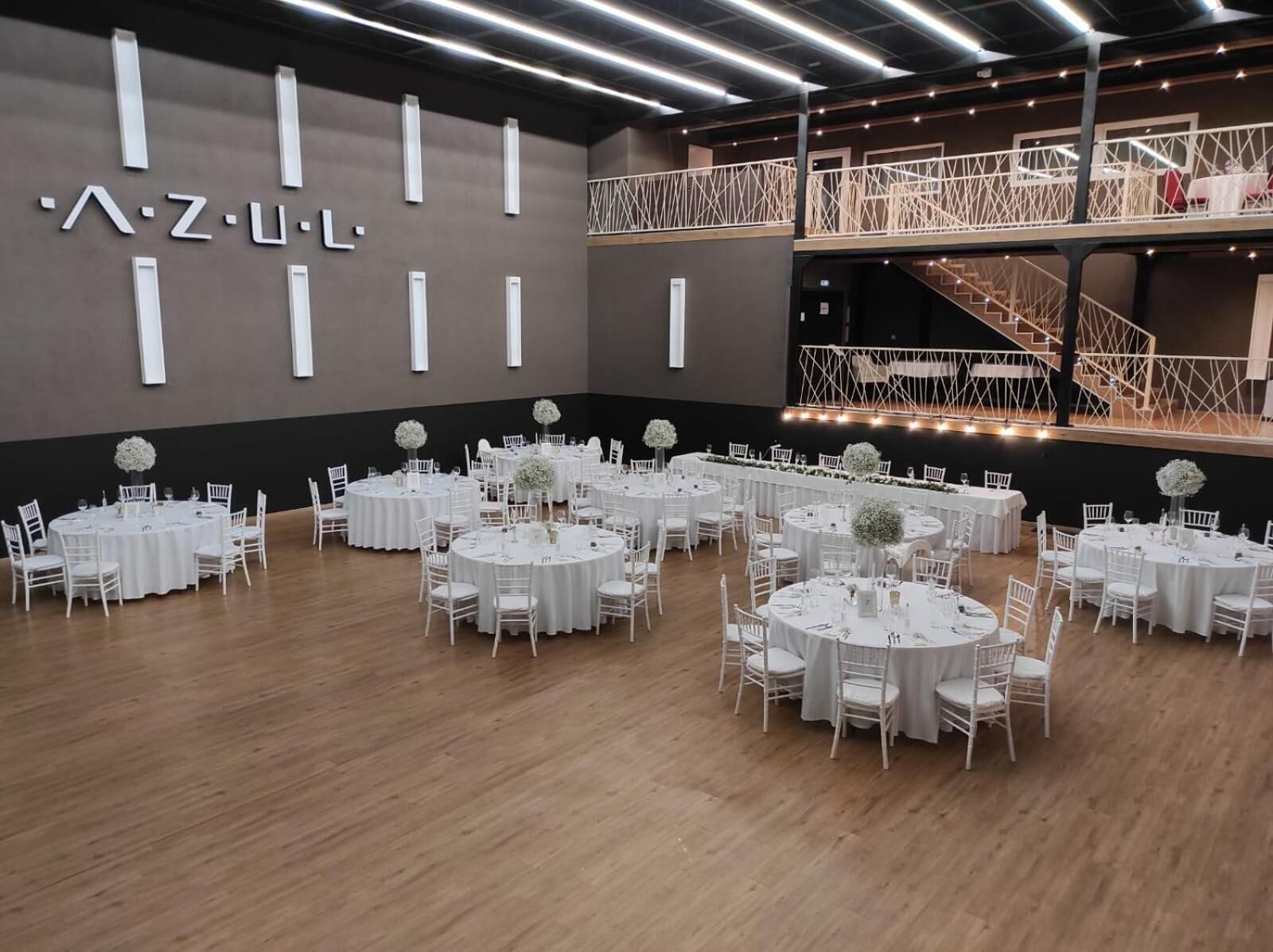 A spacious hall with multiple round tables set for events, a staircase, and a modern design with a dark wall and white accents.