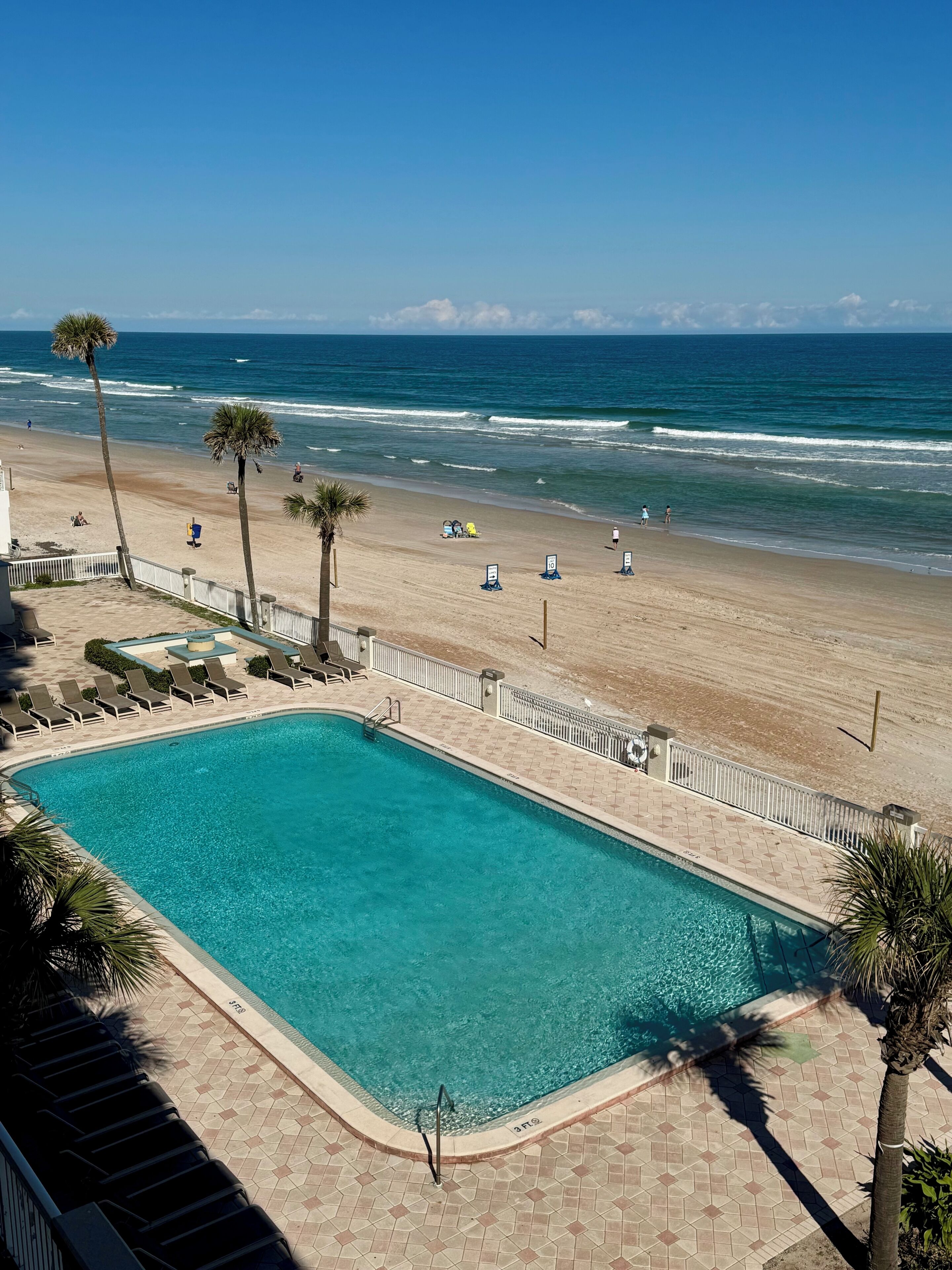 A swimming pool adjacent to a beach with palm trees and lounge chairs.