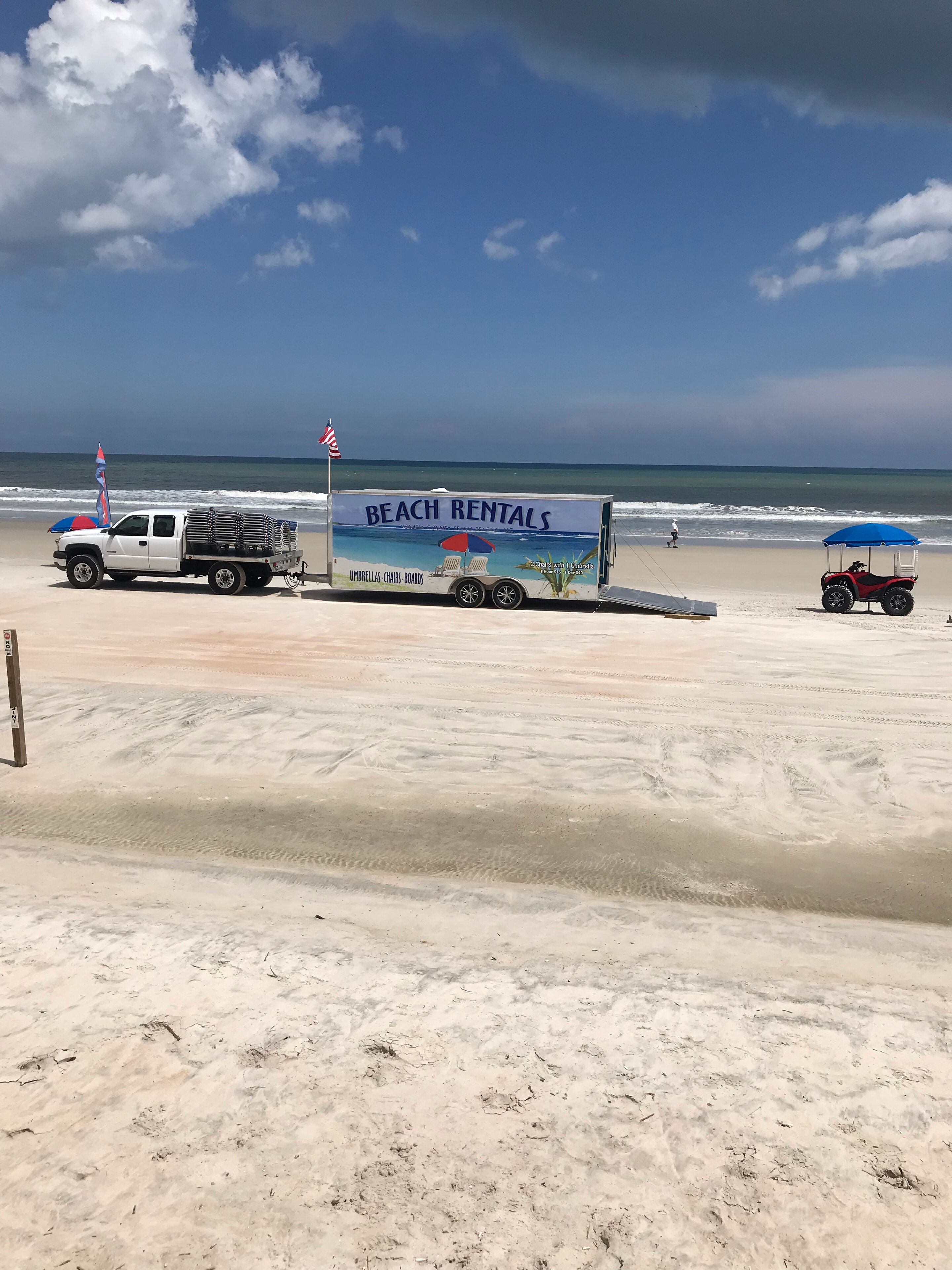 Camioneta de alquiler en la playa con un letrero publicitando alquileres de playa, una bandera y un carrito de golf sobre la arena.