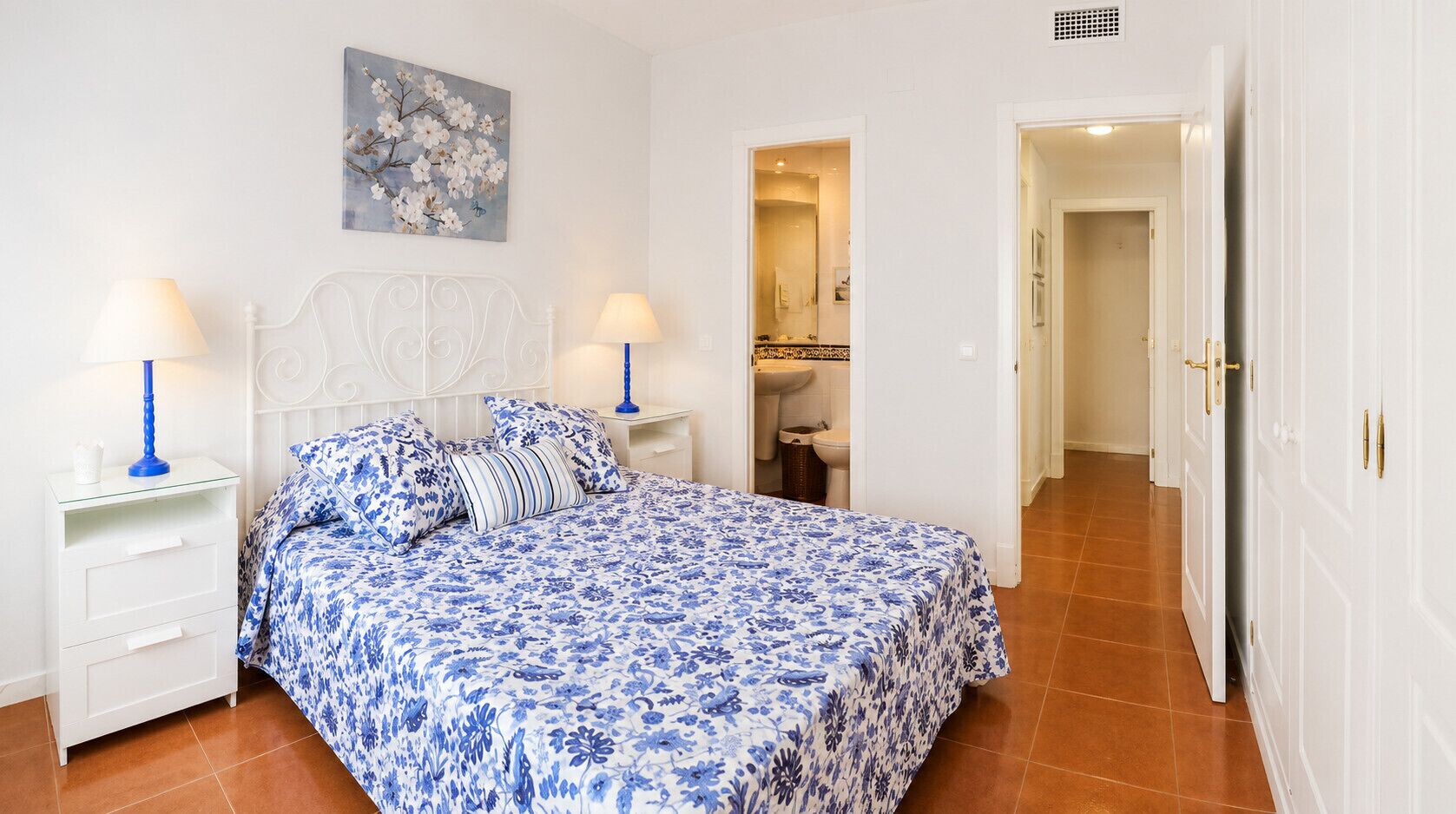 A bedroom with a bed, two bedside tables, a nightstand, a bathroom visible through an open door, and a hallway leading to another room.