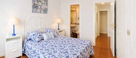 A bedroom with a bed, two bedside tables, a nightstand, a bathroom visible through an open door, and a hallway leading to another room.