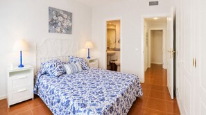 A bedroom with a bed, two bedside tables, a nightstand, a bathroom visible through an open door, and a hallway leading to another room.