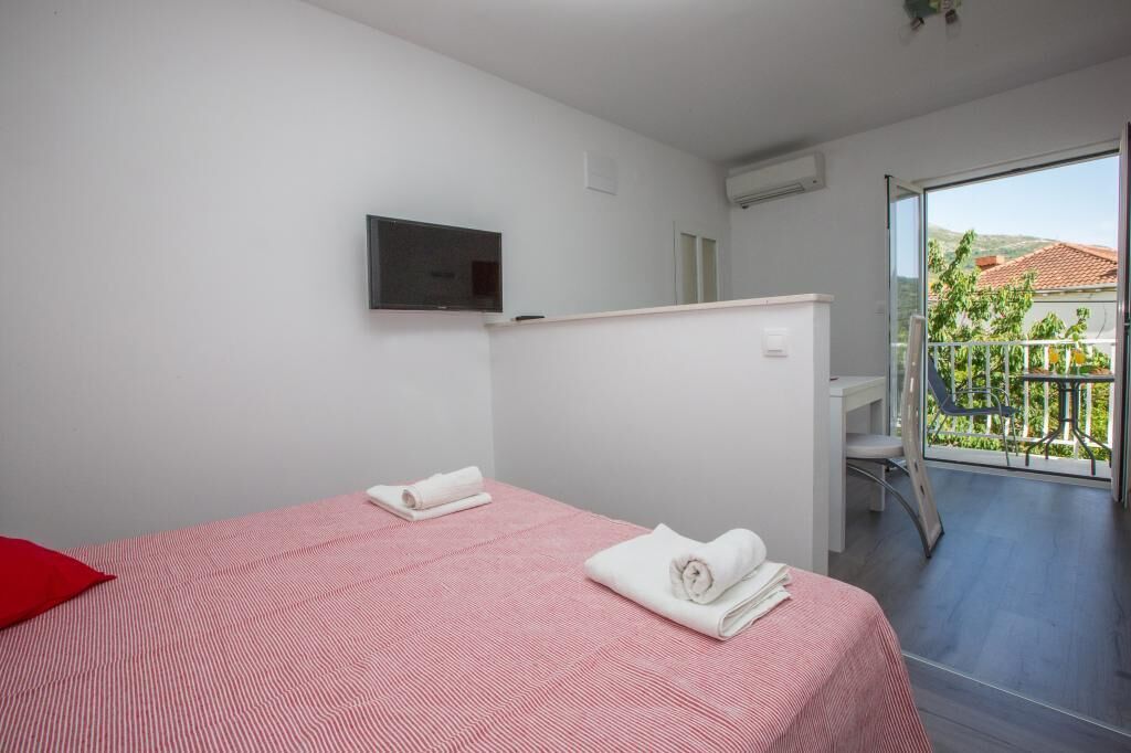 A hotel room with two beds, a TV mounted on the wall, and a balcony with a view of greenery and buildings.