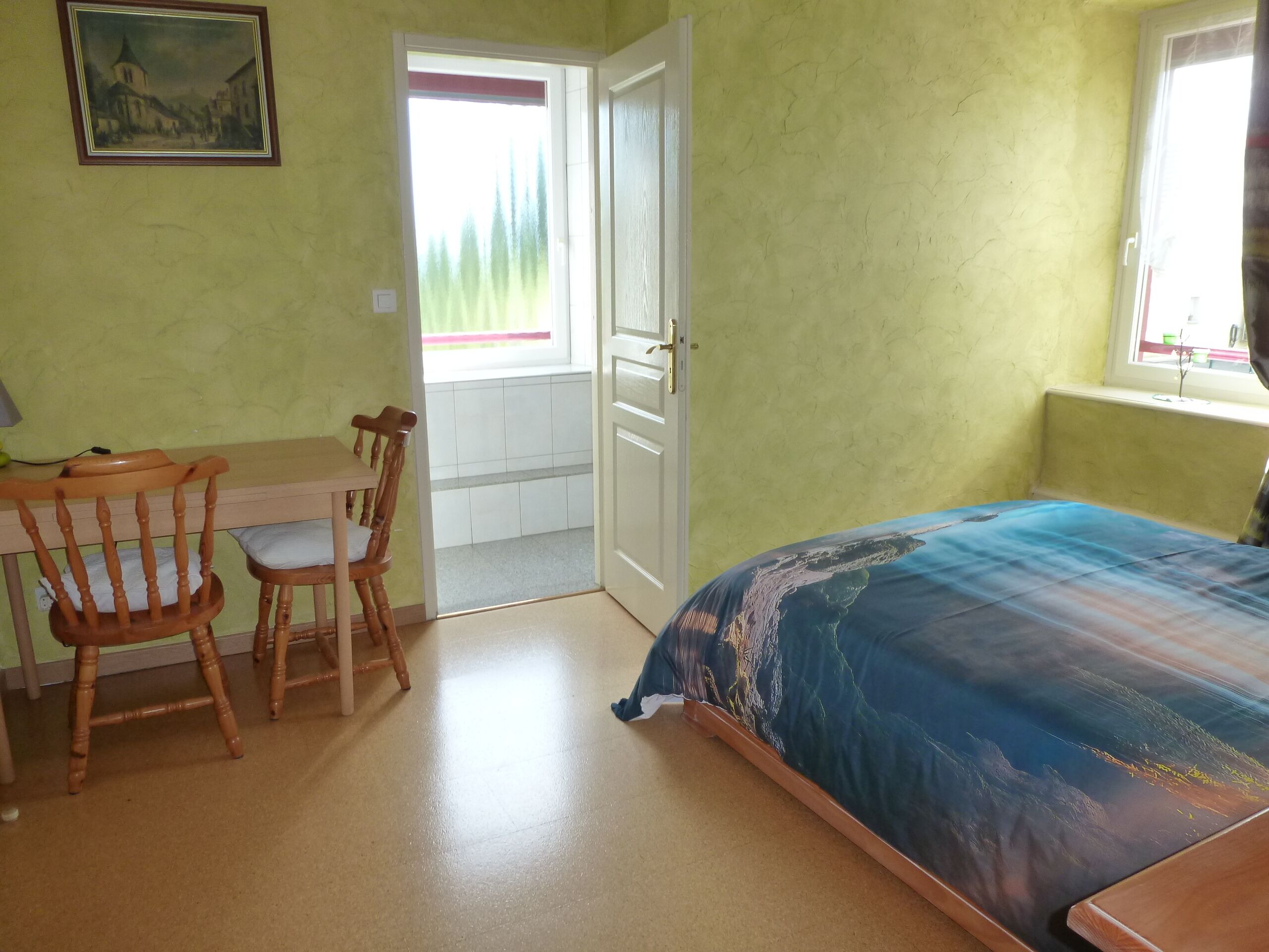 A room with a bed, two wooden chairs, a desk, a window with a view, and a door leading to an outdoor area.