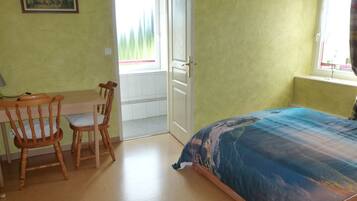 A room with a bed, two wooden chairs, a desk, a window with a view, and a door leading to an outdoor area.
