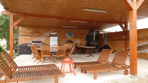 Eine überdachte Terrasse im Freien mit Holzmöbeln, einem Grill und einem Kühlschrank.