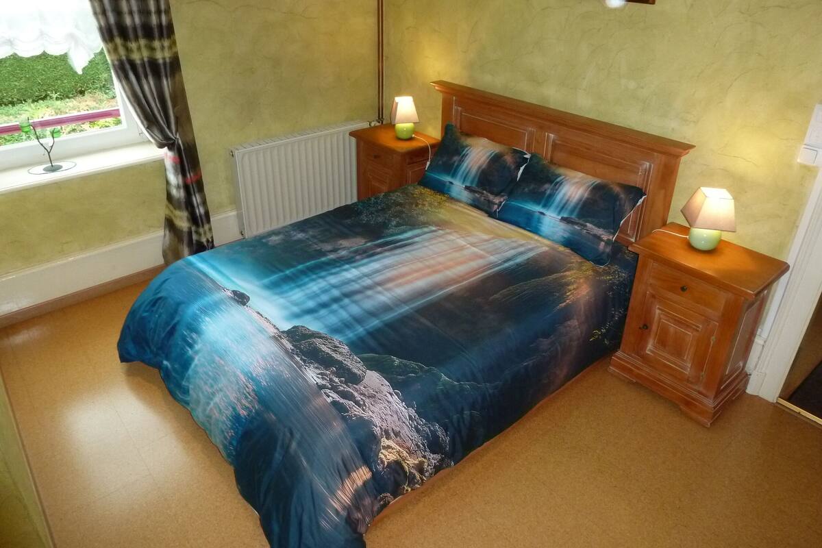 Un dormitorio con cama, mesitas de noche, una ventana con cortinas, un radiador y un cuadro colgado en la pared.