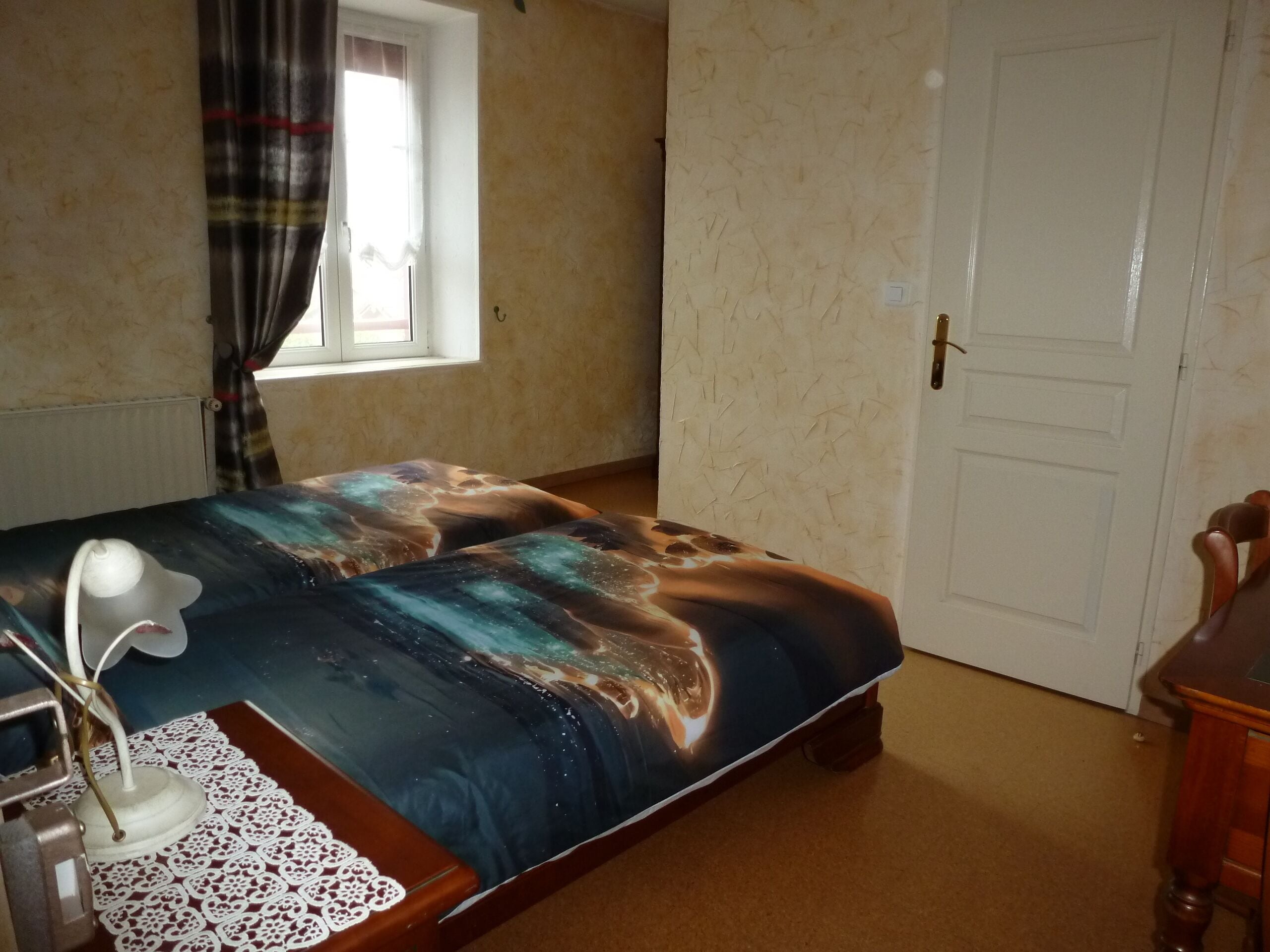 A bedroom with a bed, a desk with a lamp, a chair, a window with curtains, and a door.